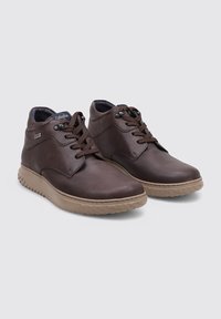 Une paire de bottines en cuir marron avec des semelles en caoutchouc beige, des bouts ronds, des laçages à l'avant et des cols rembourrés sur un fond gris clair.