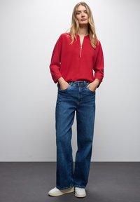 Maglione rosso con zip e spalle scese, abbinato a jeans in denim blu a vestibilità loose. Il modello indossa sneaker bianche, in piedi davanti a uno sfondo neutro.