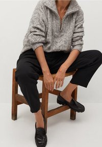 Šedý pletený svetr s polovičním zipem, volný střih a žebrované manžety. Černé zúžené kalhoty a černé nazouvací boty. Sedí na dřevěné židli.