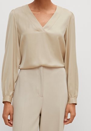 Femme portant un chemisier beige à manches longues avec un col en V, associé à un pantalon taille haute assorti sur un fond uni.