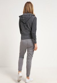 Sweatshirt à capuche grise avec un motif de crâne et du texte au dos, associé à un pantalon de survêtement gris assorti. Le matériau semble doux et texturé.