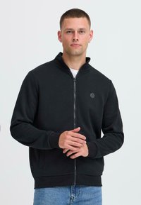 Giacca nera con zip, colletto alto, polsini e orlo a costine. Presenta un logo discreto sul petto e una superficie del tessuto liscia.