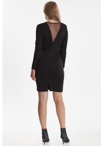 Femme portant une robe noire à manches longues avec un panneau arrière en V transparent et des bottines à paillettes noires, se tenant de dos.