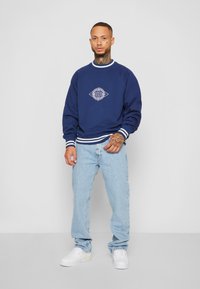 Marineblå sweatshirts med hvide accenter, prydet med et geometrisk logo, parret med lyseblå jeans og hvide sneakers. Casual outfit fremvisning.