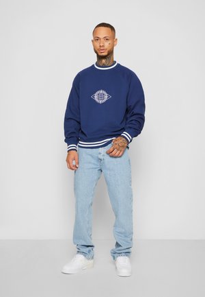 Marinblå sweatshirt med vita detaljer, med en geometrisk logotyp, i kombination med ljusblå jeans och vita sneakers. Avslappnad outfitvisning.