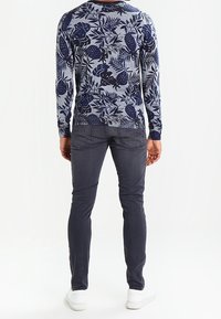 Mann in einem marineblauen Pullover mit botanischem Druck, dunklen Slim-Fit-Jeans und weißen Sneakers, der mit dem Rücken zur Kamera vor einem weißen Hintergrund steht.