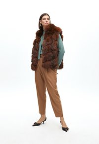Hnědá umělá kožešinová vesta přes tyrkysový top, doplněná béžovými kalhotami a černými špičatými podpatky. Vesta má texturovaný, vrstvený design.