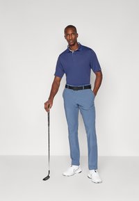 Námornícke polo tričko s krátkymi rukávmi, svetlým golierom; svetlomodré golfové nohavice; čierny opasok; biele tenisky; drží golfovú palicu.