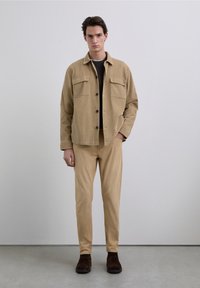 Beige fløjlsjakke med to brystlommer, knapper foran og krave, kombineret med matchende beige bukser og mørkebrune sko.