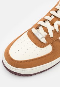 Gros plan sur l’avant d’une basket marron et crème avec des lacets blancs et des détails perforés, ornée d’une plaque métallique gravée de « "AF1" ».