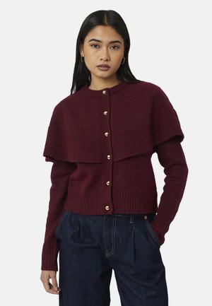 Vrouw met lang donker haar die gouden oorringen draagt en een dichtgeknoopte bordeaux gebreide cardigan over donkerblauwe spijkerbroek, staand met één hand in de zak.