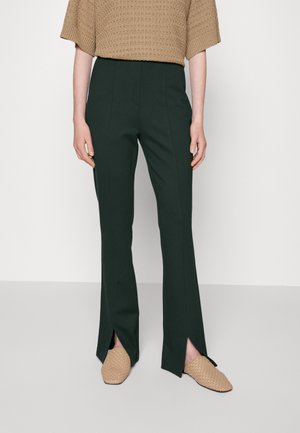 Person wearing high-waist dark green pants with front leg slits paired with beige woven slip-on shoes and a brown textured short-sleeve top.