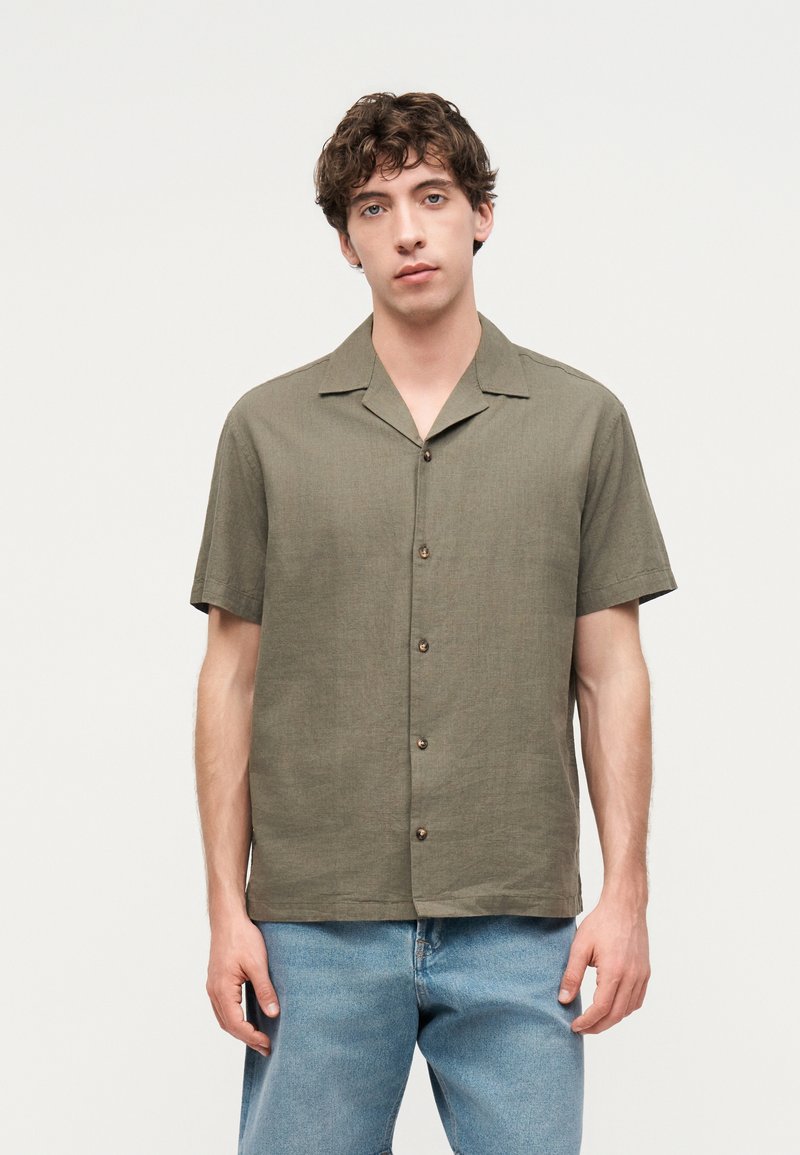 Νέος άνδρας με σγουρά μαλλιά που φοράει ελαιζωβ πήχη με κουμπιά και ανοιχτό μπλε τζιν, στέκεται μπροστά σε απλό φόντο.