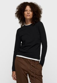 Top negro de manga larga en tela suave con cuello redondo, con un diseño sutil en relieve, superpuesto sobre una camisa blanca, combinado con pantalones marrones.