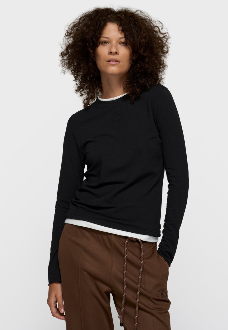 Top negro de manga larga en tela suave con cuello redondo, con un diseño sutil en relieve, superpuesto sobre una camisa blanca, combinado con pantalones marrones.