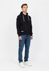 Felpa nera con coulisse bianche e testo in rilievo sulla parte anteriore, abbinata a jeans blu e sneakers bianche. Sfondo semplice.