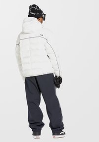 Witte pufferjas met horizontale stiksels en zwarte accenten, gecombineerd met donkergrijze losse broek en een gemêleerde zwarte beanie.