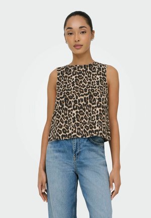 Kobieta w bezrękawniku w panterkę i jasnoniebieskich dżinsach z wysokim stanem, stojąca na tle jednolitego jasnego tła.