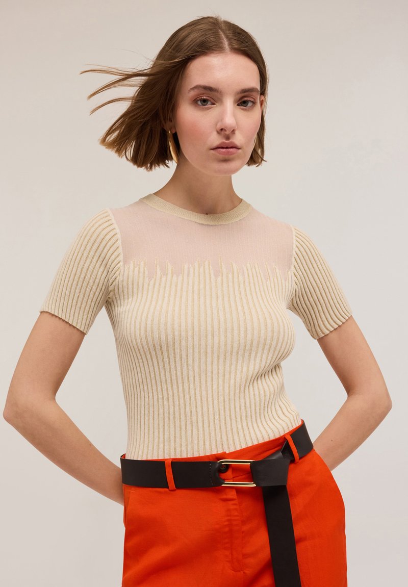 Top bej din tricot cu model în dungi, cu mâneci scurte și o secțiune superioară transparentă, asortat cu pantaloni portocalii și o curea neagră cu elemente de metal aurii.