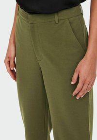 Osoba oblečená v olivově zelených kalhotách s bočními kapsami a černém topu, s uvolněnýma rukama po stranách na světlém pozadí.