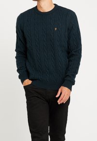 Marineblauwe kabelgebreide trui met een ronde hals, gestructureerd patroon en een klein geborduurd logo op de borst, gecombineerd met een zwarte broek.