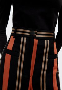 Högmidjade randiga byxor med svarta, orange och beige ränder, med platt midja och knappstängning, samt sidofickor.