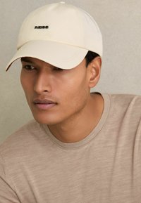 Lys beige kasket med en rund skygge, der har et sort logo på forsiden, parret med en beige T-shirt med rund hals. Glat stof overflade.
