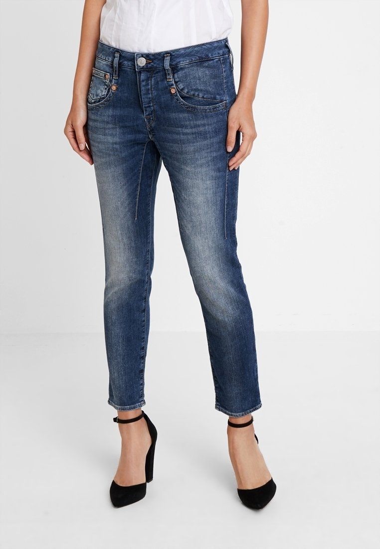 Femme portant un jean slim bleu délavé et des talons hauts noirs à bride à la cheville, debout devant un fond blanc uni.