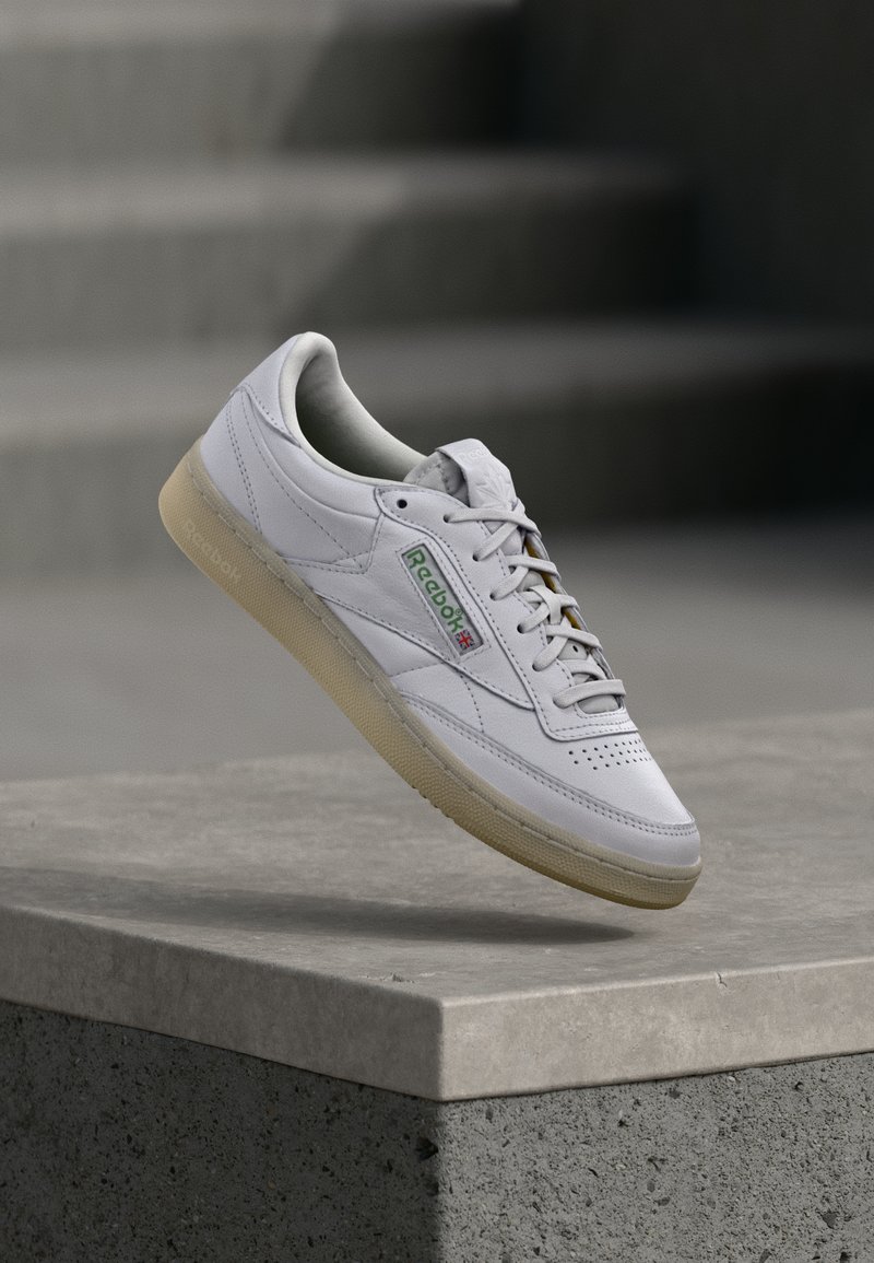White leather sneaker with a textured surface, flat laces, and green logo accents. Features a cream sole and rounded toe design.