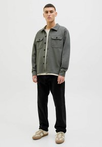 Green sweatshirt jacket with button closures and two chest pockets, worn over a cream shirt, paired with black pants and beige sneakers.