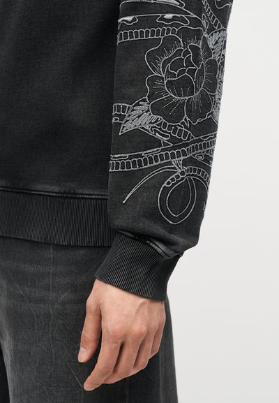 Κοντινό πλάνο μανικιού μαύρου φούτερ με λεπτομερή λευκή κεντητή λουλουδένια διακόσμηση, ριμπ μανσέτα και ένα χέρι που ακουμπά δίπλα σε σκούρο παντελόνι.