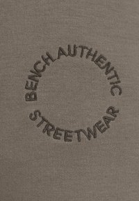 Olivgrüner Stoff mit schwarzem gesticktem kreisförmigem Text, der in fetter Schrift "BENCHAUTHENTIC STREETWEAR" lautet.