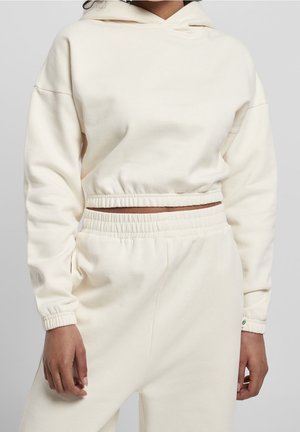 Personne portant un sweat à capuche court crème et un pantalon de survêtement taille haute assorti avec taille et poignets élastiques.