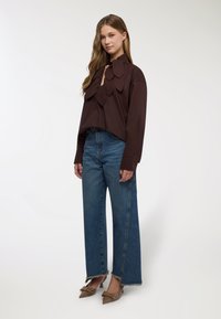 Top corto marrone con dettagli a forma di foglia, abbinato a jeans blu a vita alta e gamba larga, e scarpe con tacco marroni con decorazioni.