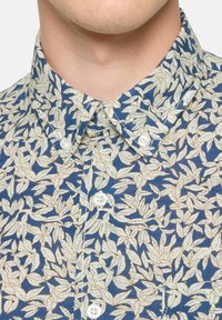 Camisa de manga corta con fondo azul, que presenta un patrón de hojas blancas, cuello mao y botones blancos. Tejido de algodón texturizado.
