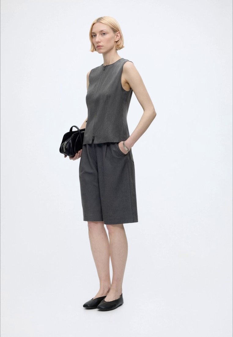 Femme aux cheveux blonds courts portant un haut gris sans manches et un short assorti longueur genoux, tenant un sac à main noir, debout devant un fond blanc.