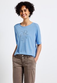 Camiseta azul claro de manga corta con escote redondo, que presenta un diseño gráfico metálico. Combinada con pantalones marrones, que muestran un ajuste casual.