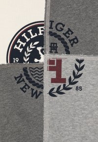 Tessuto di cotone grigio con un design a patchwork e vari loghi stampati, tra cui "HILFIGER" e motivi di alloro in blu navy e bordeaux.