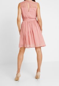 Robe rose sans manches avec des détails en dentelle dans le dos, taille cintrée, et une jupe évasée arrivant juste au-dessus des genoux.