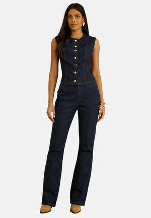 Vrouw draagt een donkerblauw mouwloos denimtopje met gouden knopen en bijpassende hooggetailleerde denimbroek, beige puntige schoenen en gouden sieraden.