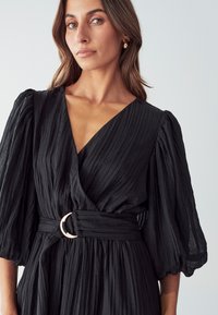 Black dress with pleated texture, deep V-neck, wide puff sleeves, cinched waist featuring a gold circular buckle detail.