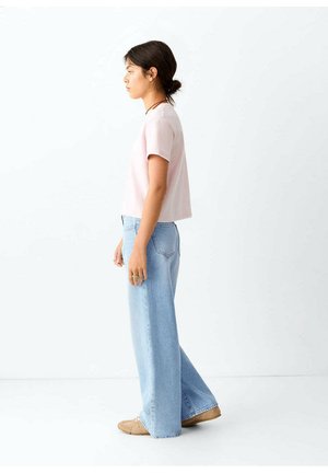 Mujer joven de perfil que lleva una camiseta de manga corta rosa claro, vaqueros anchos azul claro y zapatos marrones sobre un fondo blanco.