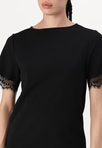 Svart topp med korte ermer og intrikate svarte blonder på kanten. Glatt stofftekstur og design med båtutringning.