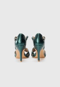 Teal leather high heels with a glossy finish, open back, and crisscross straps. Featuring a slender stiletto heel and beige sole.