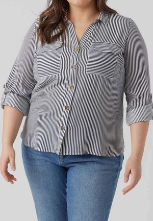 Femme portant une chemise à rayures verticales noires et blanches avec des poches poitrine et des manches retroussées, associée à un jean bleu.