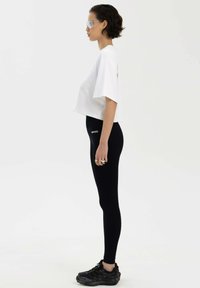 Biała skrócona koszulka, czarne legginsy z logo i czarne buty sportowe. Modelka nosi przezroczyste okulary przeciwsłoneczne. Prosty design i dopasowany kształt.