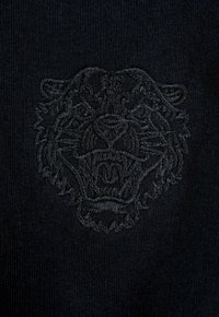 Czarna dzianina z wypukłym haftowanym wizerunkiem lwa na środku, z bogatymi detalami i gładką teksturą.