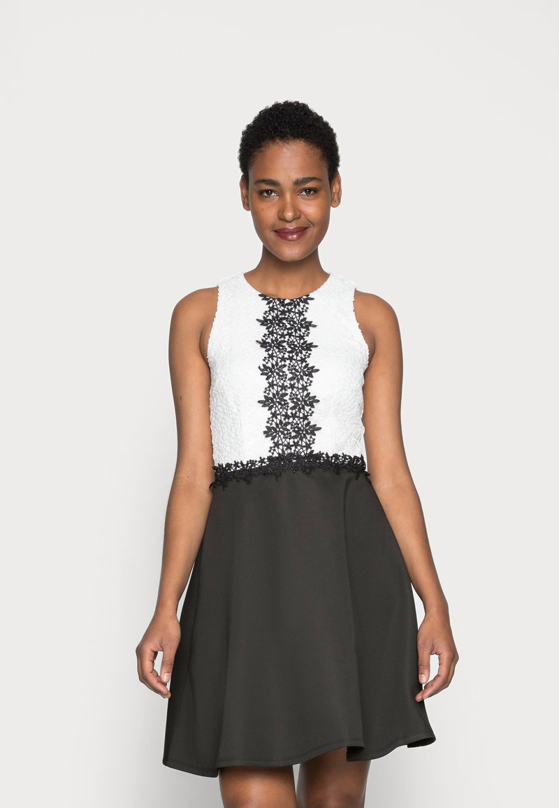 Schwarz-weißes A-Linien-Kleid mit einem strukturierten weißen Oberteil, aufwendigen schwarzen Spitzenverzierungen und einem glatten schwarzen Rock.