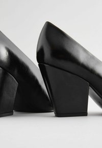 Svart lackläder högklackade skor med geometrisk kilklack. Slät, glänsande yta med spetstoe-design. Sidsyn som framhäver formen.