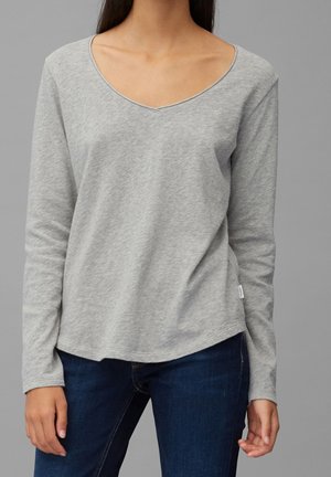 Vrouw met een lichtgrijs langemouwen shirt met V-hals en donkerblauwe spijkerbroek tegen een effen grijze achtergrond.